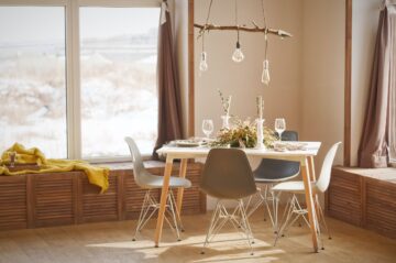 The image shows a naturally lighted room with a set table and four chairs around it. There are benches directly in front of the windows and a blanket on one of them. There’s a hanging light fixture above the table.