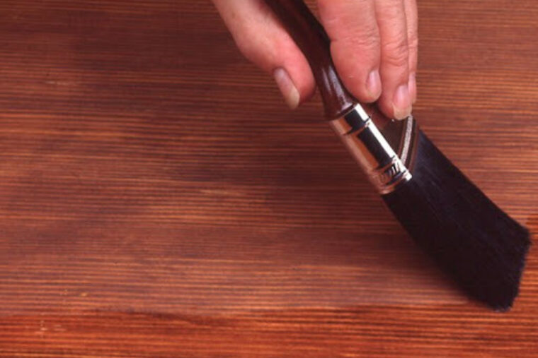 Person is staining a piece of wood using a wooden paintbrush with black bristles.