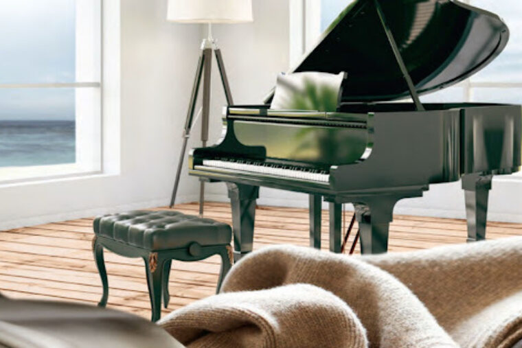 This living room has a sofa with a blanket laying across it and a green piano and piano bench placed in the middle of the room overlooking the view of the ocean.