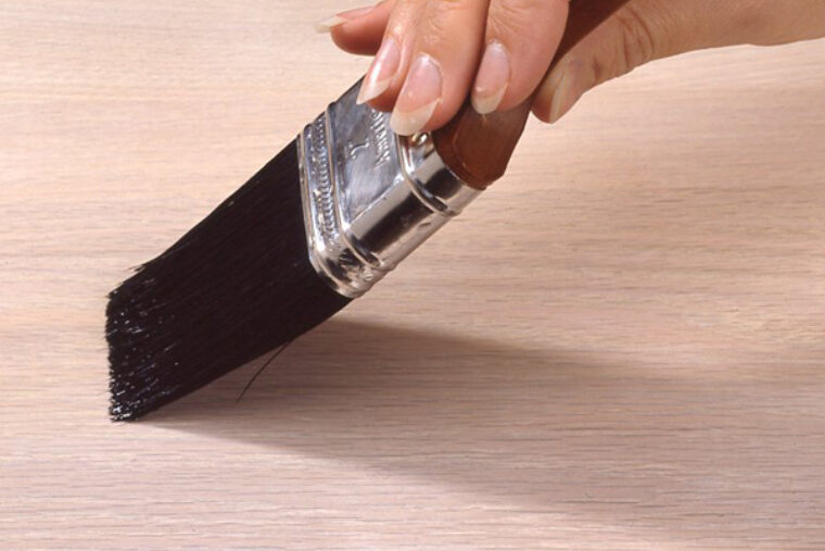 A person is staining a piece of wood with a wood-handled paintbrush with black bristles.