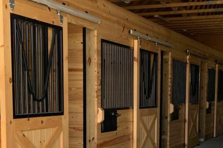 This horse stable has sliding doors with black bars. There are wooden rafters, along with concrete flooring.