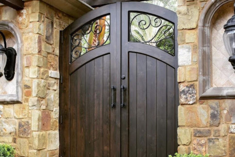 A beautiful wooden door with black handles is standing between tan, stoned walls. There are shrubs along the side of the entryway.