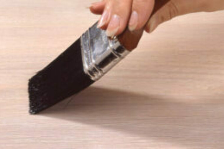 Someone is applying polyurethane with a brush, The brush has black bristles and a wooden handle.