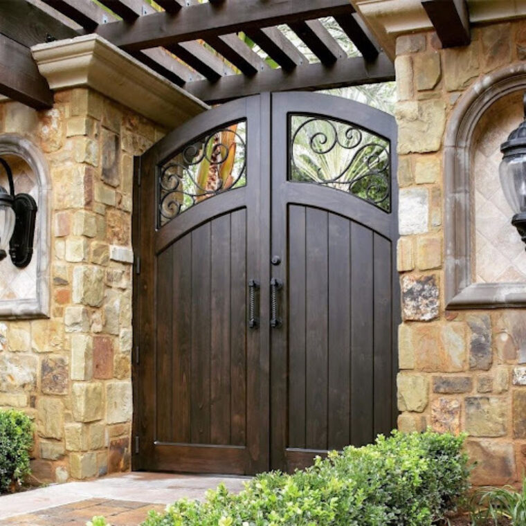 A beautiful wooden door with black handles is standing between tan, stoned walls. There are shrubs along the side of the entryway.